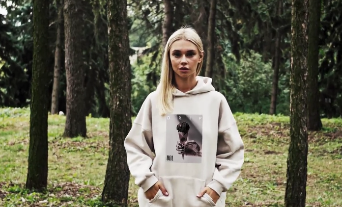 A person stands in a forest wearing a light hoodie with a graphic print, surrounded by trees and greenery.