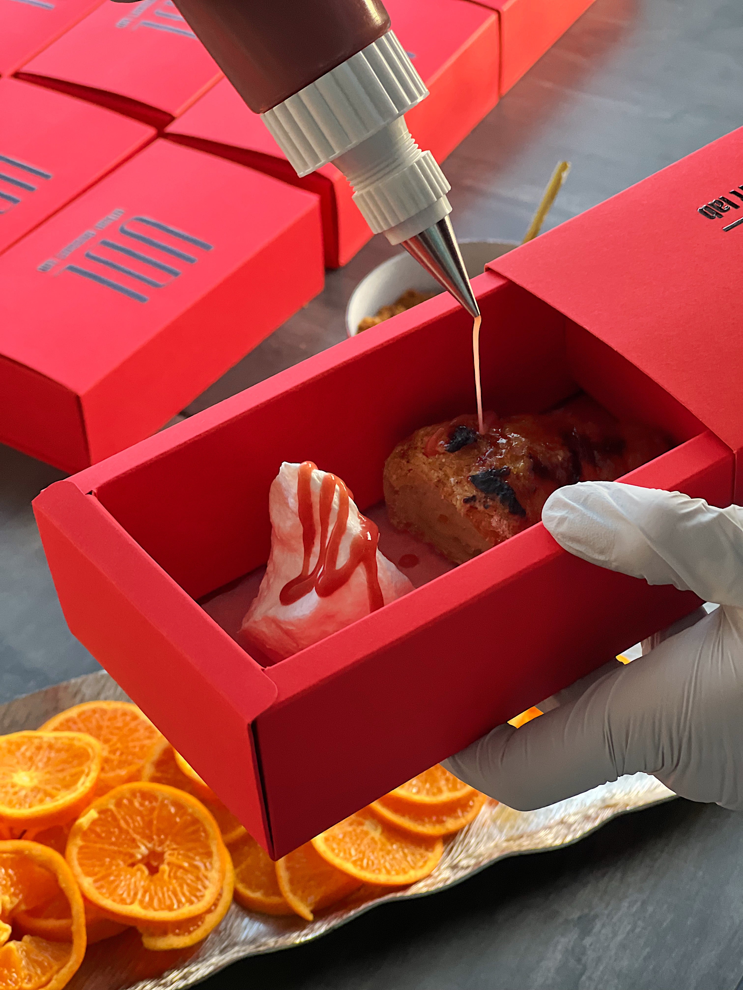 A hand decorating a croissant pastry with sauce in a Branded Red Urban Dessert Lab boxes. Orange slices on a tray and a bowl of dessert toppings are in the background.
