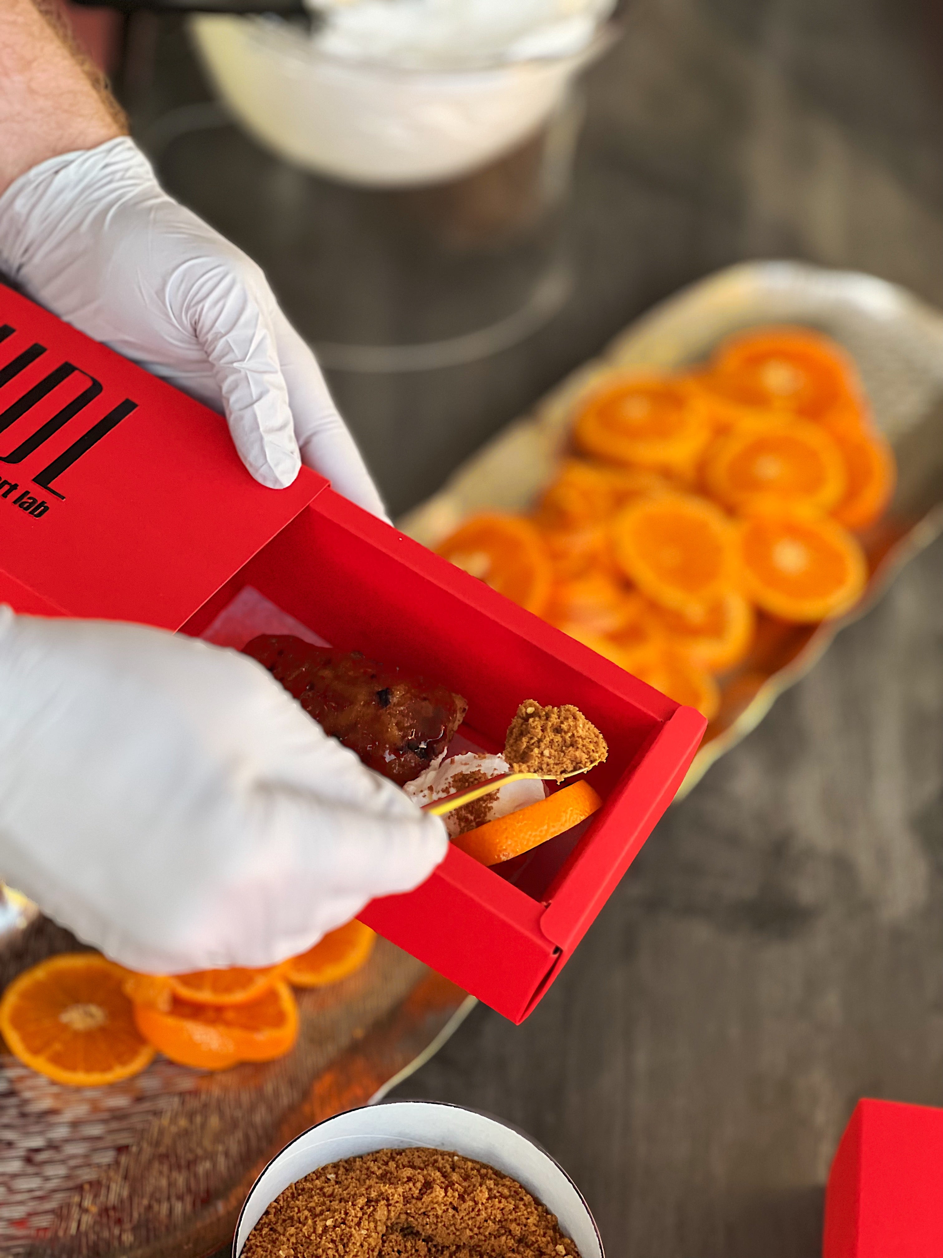 A hand decorating a croissant pastry with dessert crumble in a Branded Red Urban Dessert Lab boxes. Orange slices on a tray and a bowl of dessert toppings are in the background.