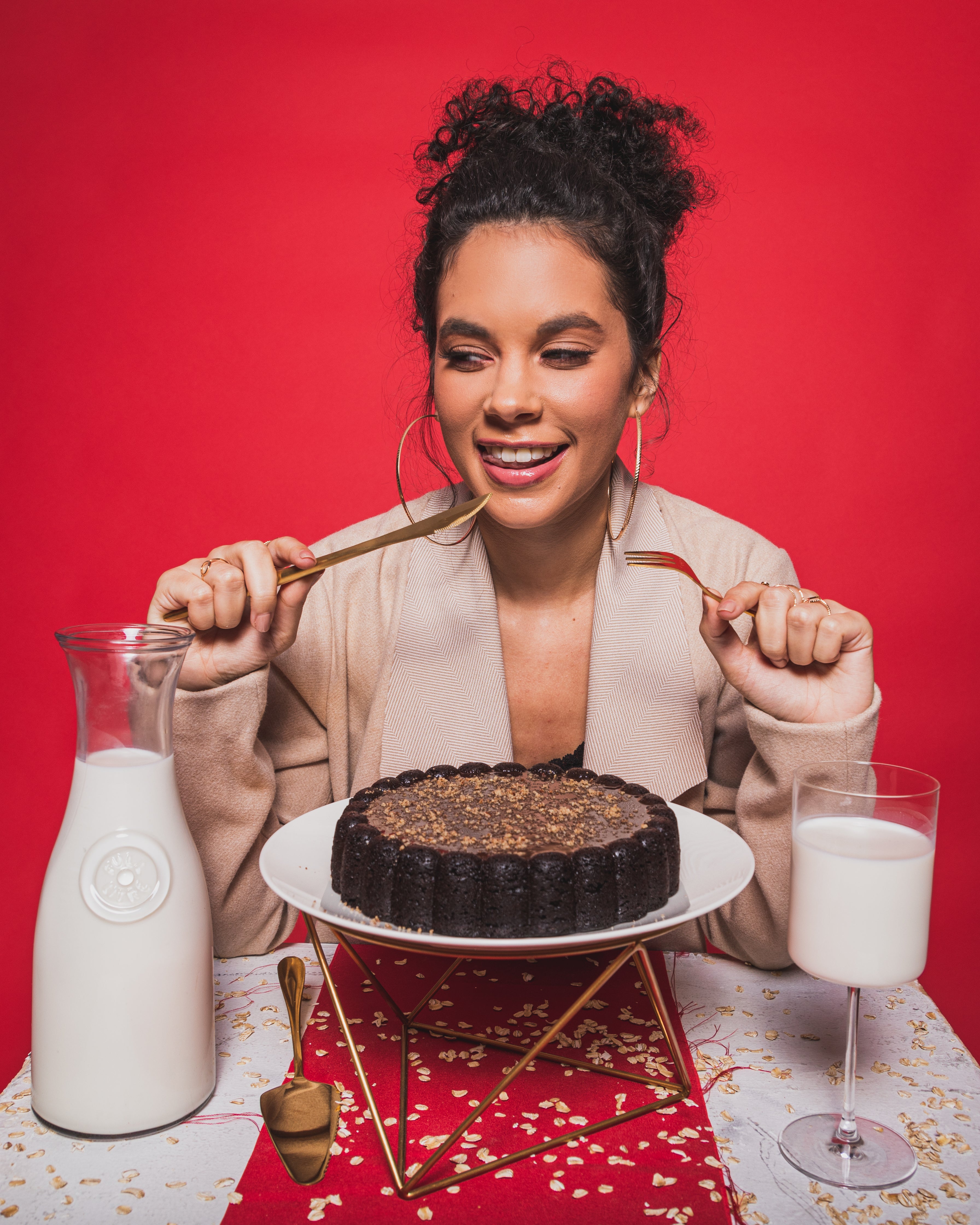 model holding cutlery in front of a chocolate cake on a cake stand. A jug and glass of oat milk are also on the table.