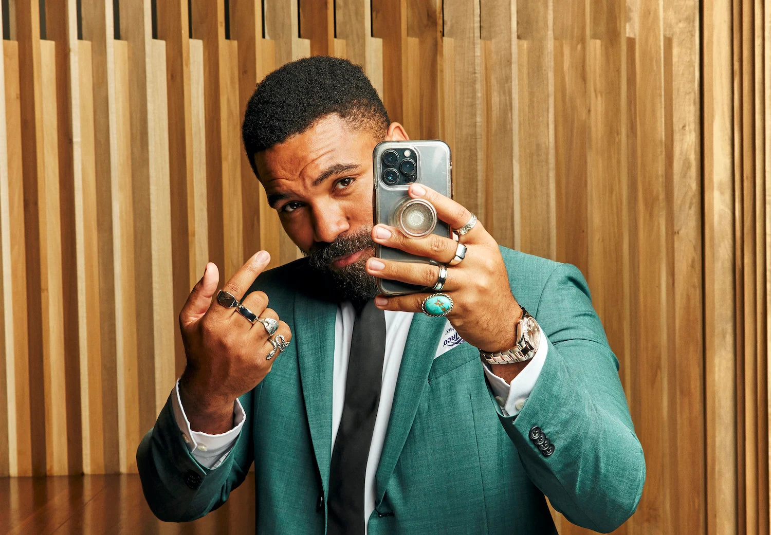 Jordan Stallion, a popular Influencer poses for a selfie, showcasing multiple rings and a stylish watch, with a wooden backdrop background