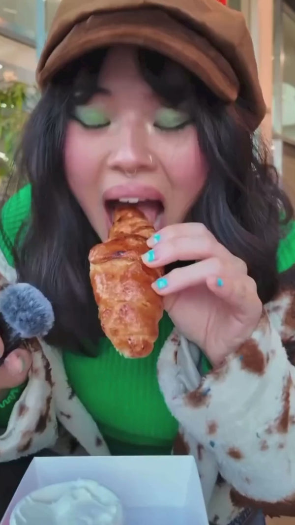 A person with dark hair and green eyeshadow is about to eating an Urban Dessert Lab bruleed croissant while holding a microphone.