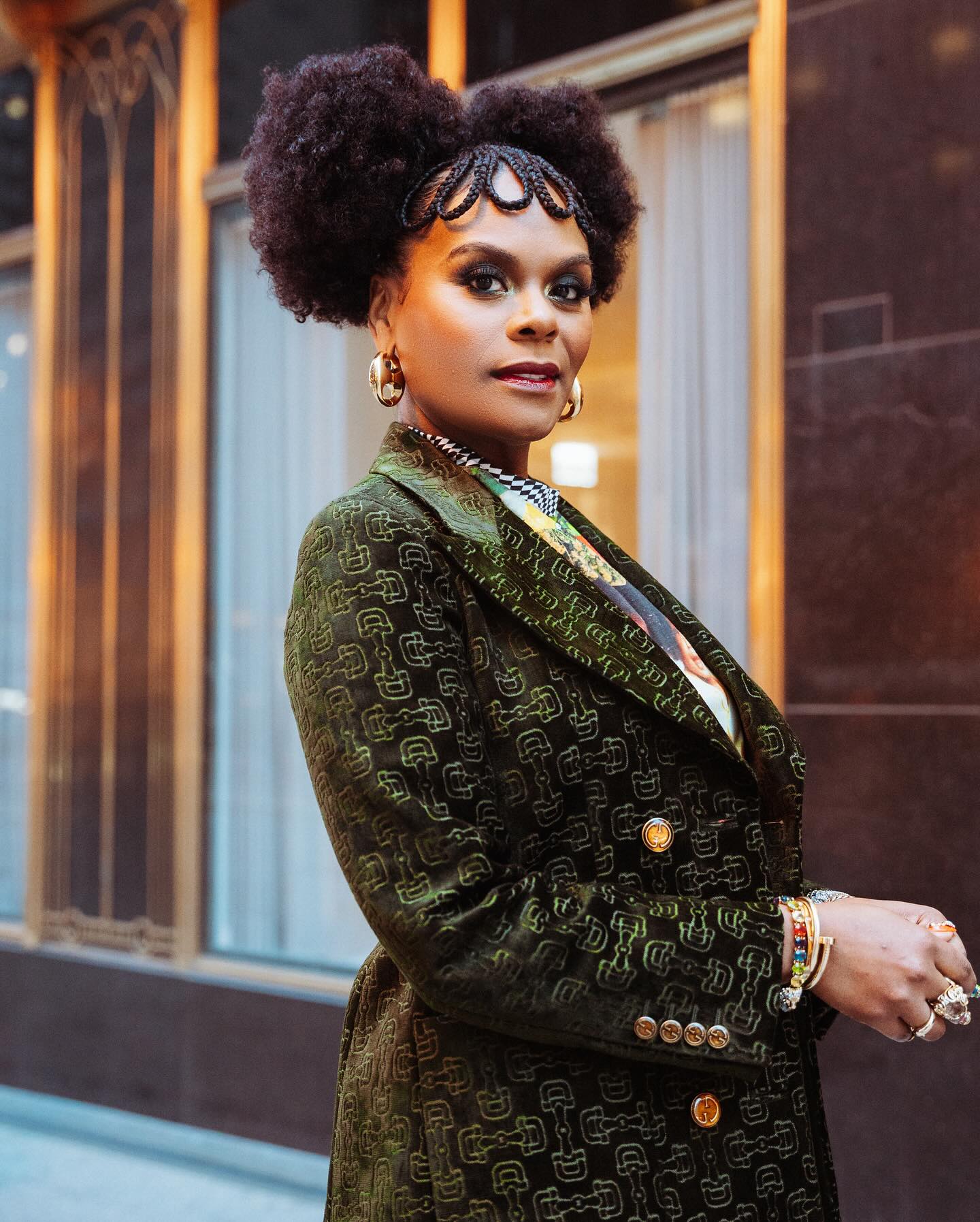 Tabitha Brown, a stylish individual poses outdoors, wearing a green patterned blazer and statement jewelry, with curly hair styled in an elegant updo.